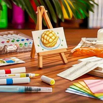 A small wooden easel holding a painted drawing of a golden pastry on a plate, surrounded by art supplies including markers, watercolor paints, paper sketches, and colored pencils on a wooden table.